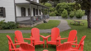 A ring of eight red muskoka chairs are joined at the arms creating an enclosed area, presented on a green turf lawn in front of an older home.