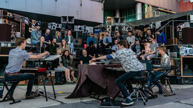 musicians play synths to a crowd seated on arena-style bleachers assembled from 400 loud speakers