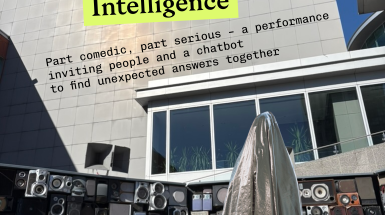 a person cloaked in a silver fabric, resembling a ghost, stands atop a sculpture made of old speakers
