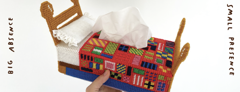 a hand holding a cross stitched tissue box that looks like a bed with text on either side of the image that reads "Big Absence" and "Small Presence" in handwritten text