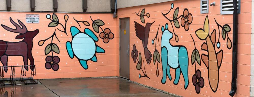 Mural with colour pallet that uses orange, turquoise, and browns. Mural depicts different wild life and plants. Such as a caribou, bear, turtle, humming bird, and fish.  