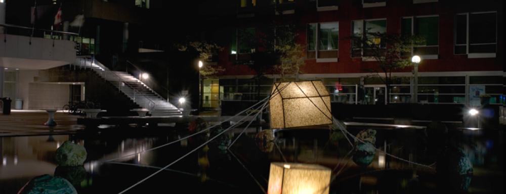 A glowing sculpture of house suspended on water tied to large rocks with rope.