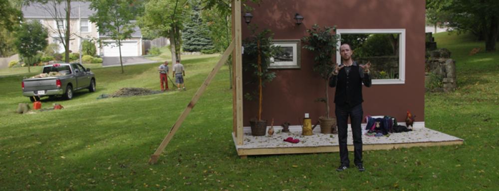 A structure made too look like a room with only two walls. Placed in a park. With man speaking in front of it. 