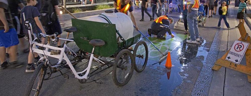 A two seater bike with wagon that holds a water tank. Two men cleaning the bike route sign painted on the ground. 