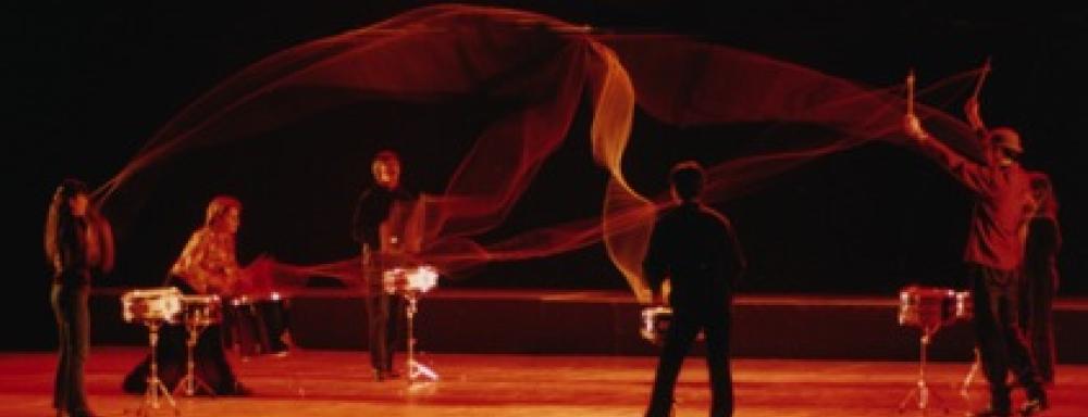 Six individuals playing drums with nylon rope attaching their sticks to other's.