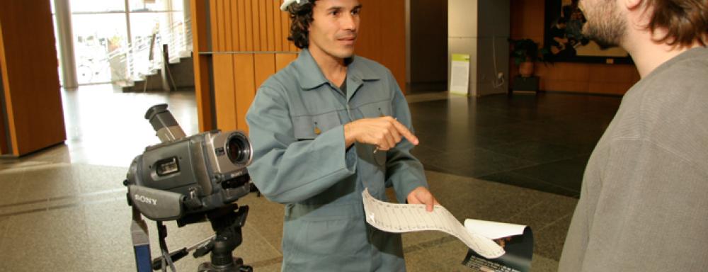 Man in construction hat behind a video camera pointing at a paper talking to another man. 
