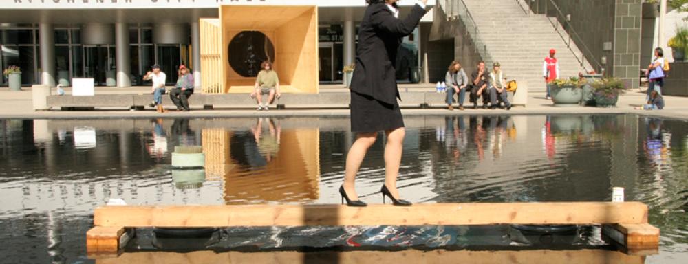 A individual standing a top a wooden balance beam in the middle of water. Wearing heals and pantsuit.
