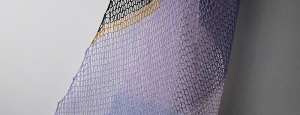 Knitted yarn hung in the shape of a sail boats sail. The yarn is purple, black, white and gold. 