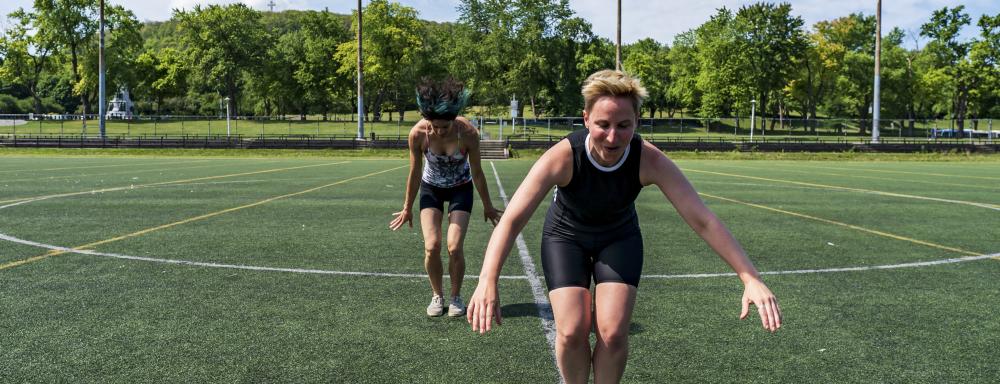 Two women in a sports field with feet together and arms out for balance, hair flying, as though they are jumping with both feet