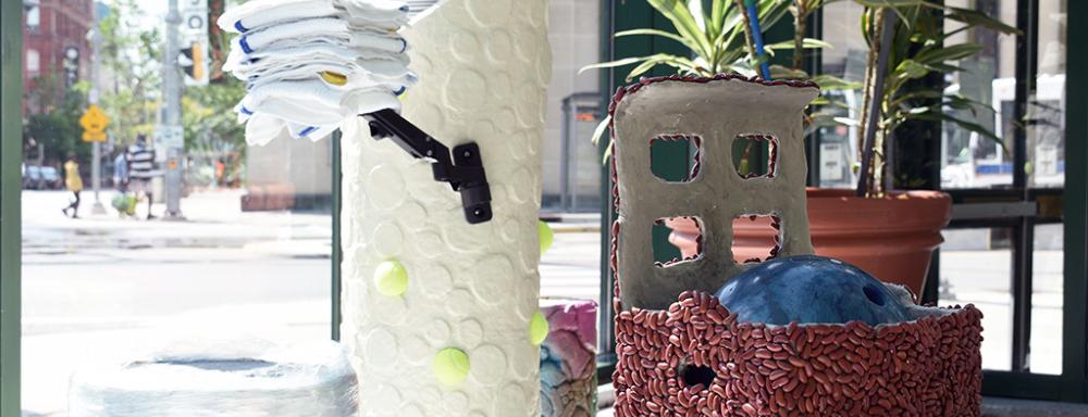 A series of sculptures - one is a box covered in kidney beans holding a bowling ball, another is a white-polka-dot tower filled with stacked plates, with an arm holding towels, and some of the polka-dots are replaced with tennis balls.