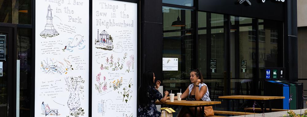 Two people sitting at a table outside cafe pyrus in front of a window vinyl depicting sketches and descriptions of locations or sights in the neighbourhood