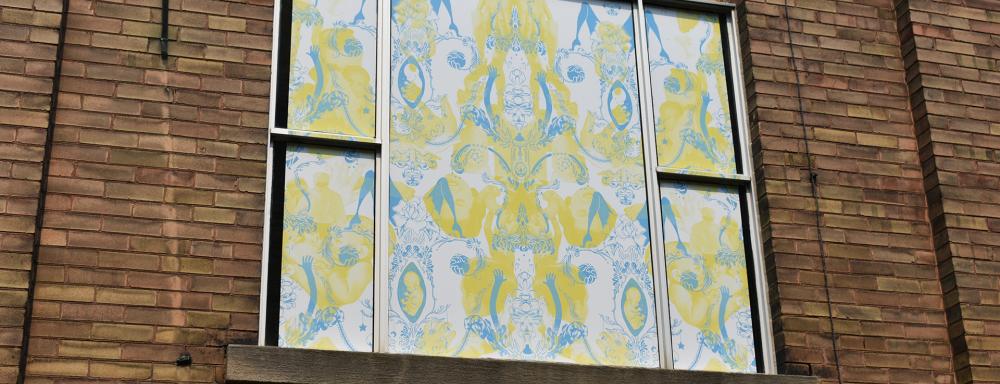 A window in a brick wall-papered over by a yellow and blue pattern on white background. The yellow pattern has images of male and female adult heads, and the overlapping blue has a floating fetus, spread legs, and a floral theme.