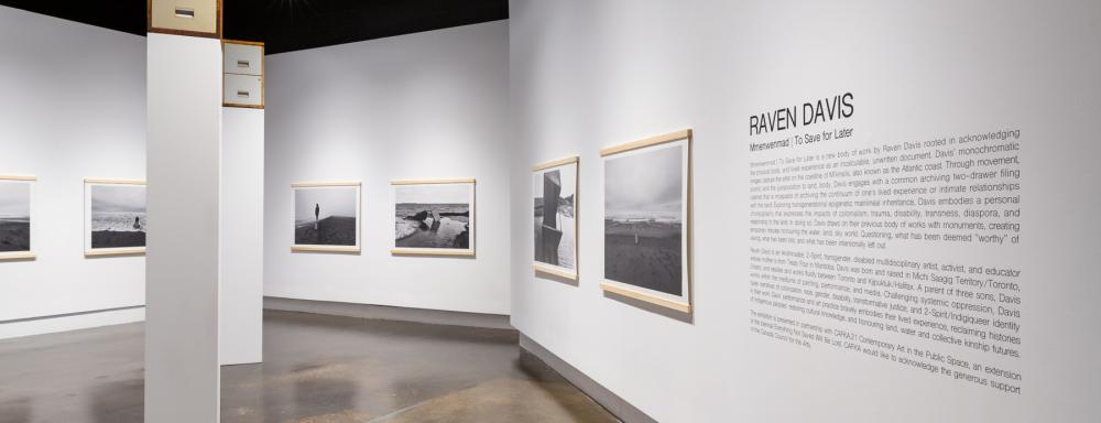 White walls of an art gallery, with black and white photographs framed only on the top and bottom with wood. There are also two double drawer filing cabinets on top of plinths far above head height.