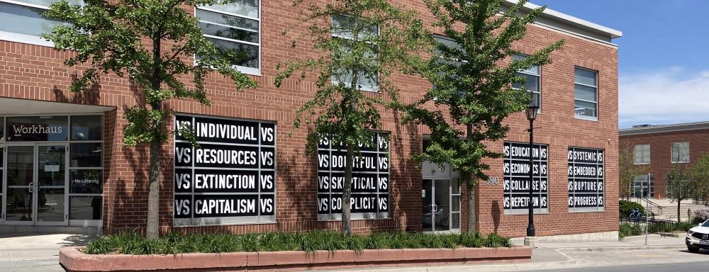 A view of a brick building with windows blacked out, with white text that says in rows "vs individual vs" "vs resources vs" "vs extinction vs" "vs capitalism vs" and many more similar phrases