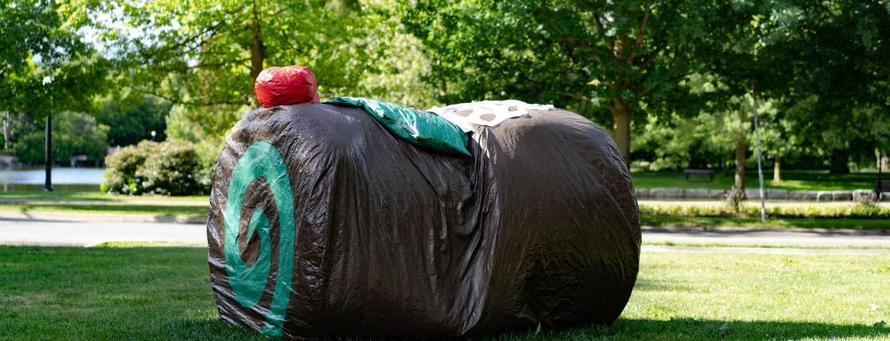 A brown cloth material in the shape of a cylinder on its side, perhaps 5 feet in height. There is a minty green swirl on the circular end visible, and a cloth cherry and green cloth leaf on top.