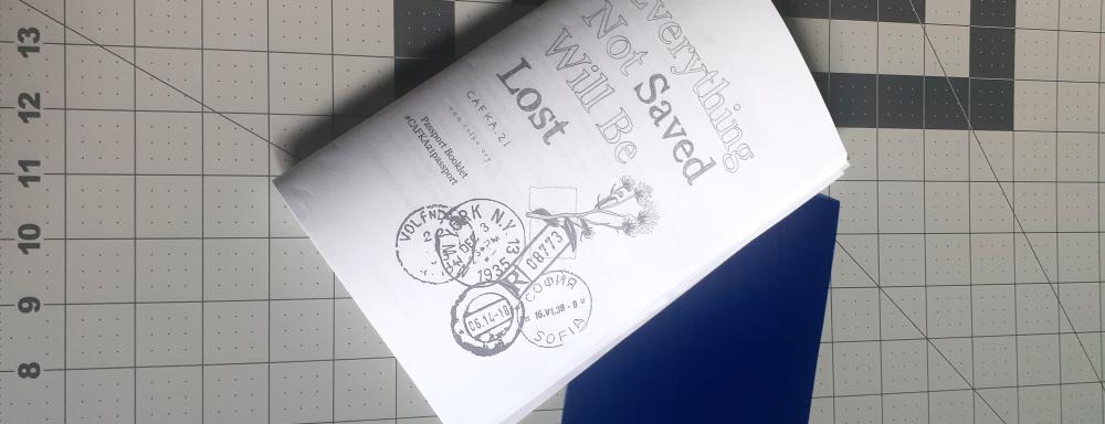 A folded paper booklet sits on a line ruled cutting mat. The cover has the title Everything Not Saved Will Be Lost and several passport stamps