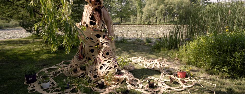 A person standing in a green area wearing a dress with hood composed of woven rope, holding a variety of potted plants down the body of the dress and in the expansive trail of weaving spread onto the ground.
