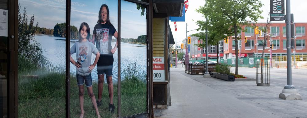 A vinyl print of a photograph depicting two Indigenous people standing in front of green plants and a lake with their fists held to hips and elbows out. The right figure is an indigenous adult, in a black t-shirt with an old newspaper article titled "Indian Land for Sale" but "for sale" is crossed out with red and replaced with "not for sale". On the left is a youth wearing a t-shirt picturing a red-brown skinned angry hulk behind an indigenous man reading "treaty broken". Installed on a shop window.