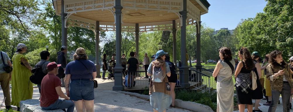 Many people standing around a gazebo to listen to a 5-person choir perform, sunny day