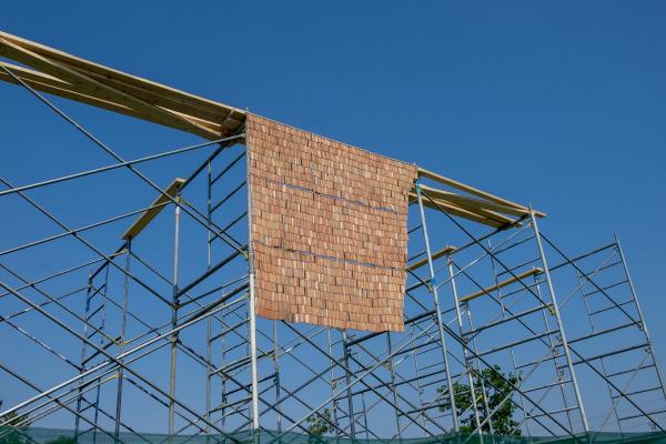 Looking upward at a terracotta-coloured curtain made from thousands of handmade beads hangs from scaffolding with a clear blue sky in the background.