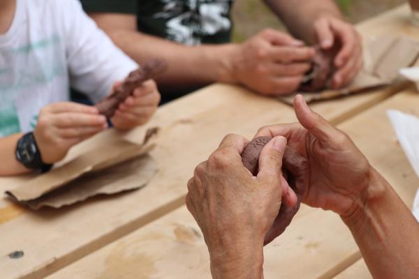 Three sets of hands, sitting around a wooden picnic table working with brown coloured clay