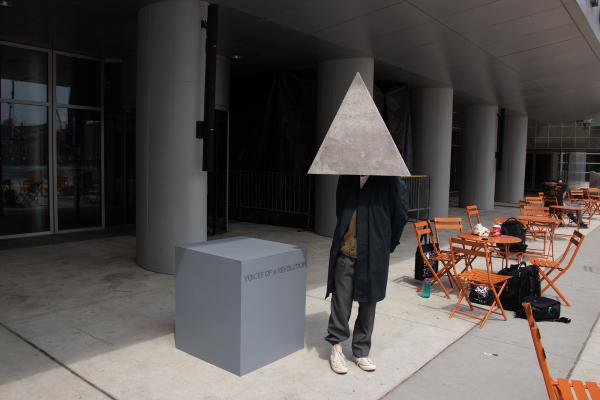 Person in white sneakers, a long navy coat, wearing a grey, textured, triangular helmet that obscures their face, standing beside a grey plith with text that reads "VOICES OF A REVOLUTION"