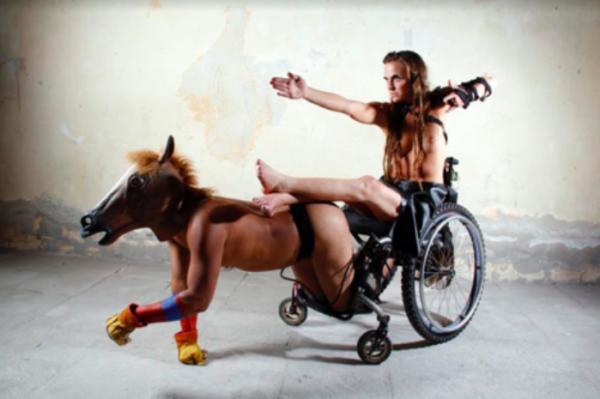 Image of man in wheel chair being pulled by man on all fours in horse mask