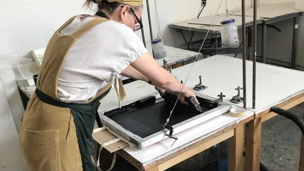 Person in beige overalls standing over a table, pulling black ink through a silkscreen.