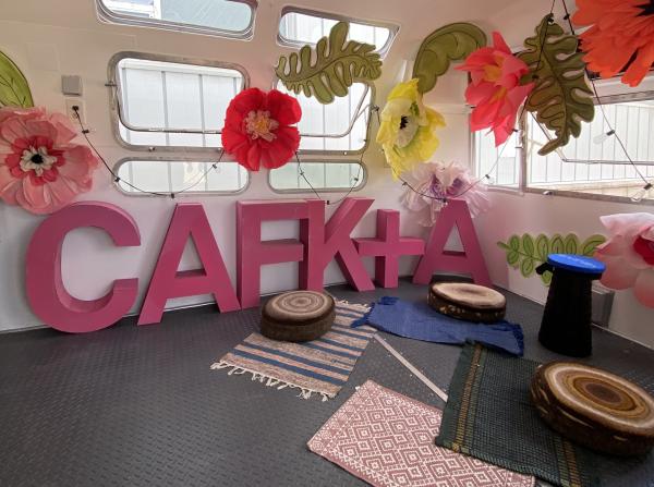 The CAFKA logo made in large pink cardboard letters, set against a wall with windows surrounded by oversized paper flowers. Next to the CAFKA letters is a collection of floor seating cushions
