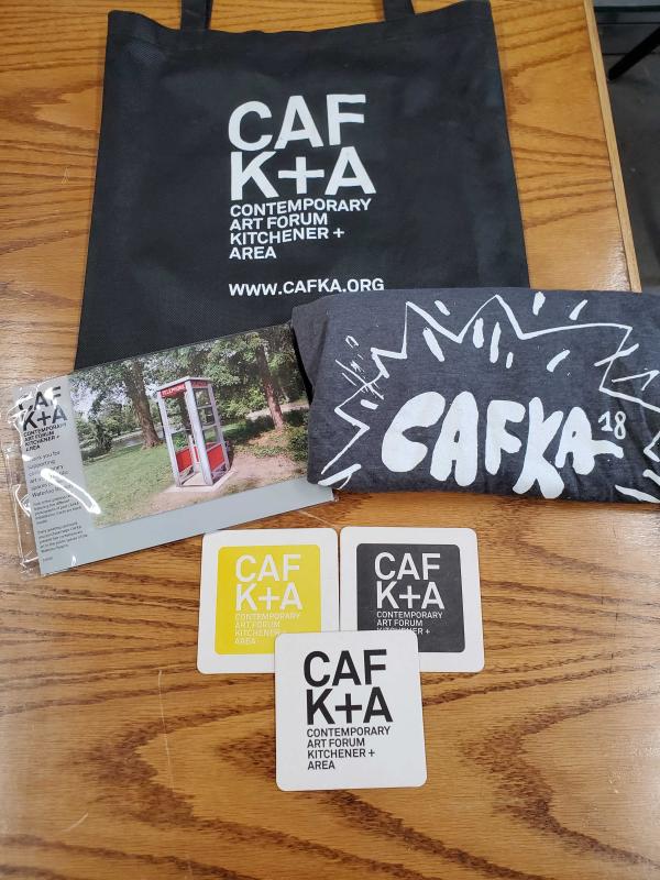 A collection of items sits on a table, including a black tote bag with the CAFKA logo in white, a set of postcards featuring previous biennial artworks, a grey CAFKA 18 t-shirt, and three CAFKA logo coasters
