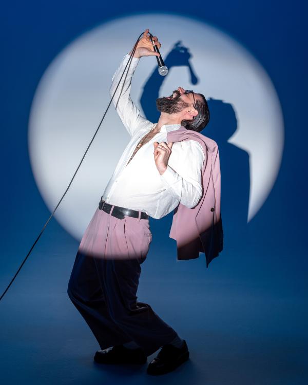 Ben Gorodetsky in a spotlight, wearing pink suit pants and holding a suitjacket over his shoulder. With the other hand, Ben dangles a microphone over his mouth, head tilted back.