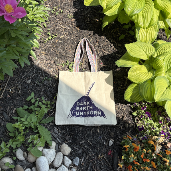 A tote back with a rainbow striped handle lies in a garden bed, surrounded by rocks and flowers. The tote bag, which is natural canvas, shows an indigo screen print of a dirt pile with a unicorn horn poking out. The dirt pile reads "D4RK E4RTH UN1KORN".