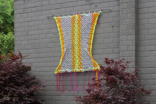 Tapestry hanging on a grey painted brick wall. The tapestry resembles chainlink fence – silver-grey, orange, and yellow ribbons are woven, into striped panels. Ribbons cross-over in a diamond shape. Hot pink ribbons hang from the bottom of the piece like tassels.