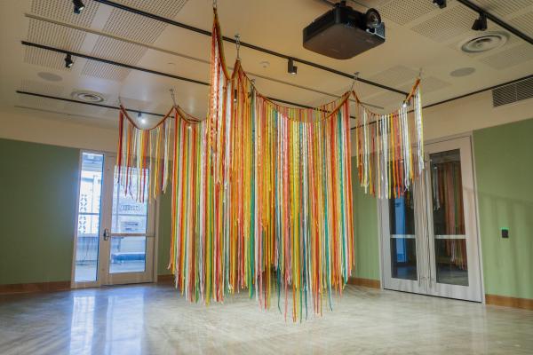 Pink, green, yellow, red, and white ribbons hang together in a tent-line piece, suspended from the ceiling in a room with green walls.