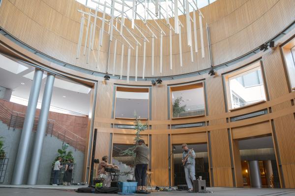 Three people making music with a blue atlas cedar tree in a round room. From the ceiling, a sculpture hangs in the shape of the wooden skeleton of a house.